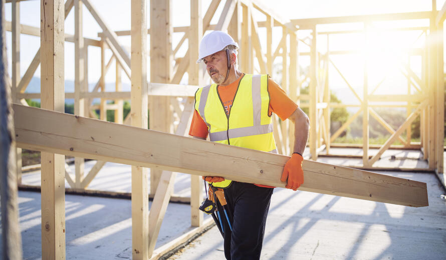 Side view of builder holding wooden beam, working on unfinished wooden house at sunset.