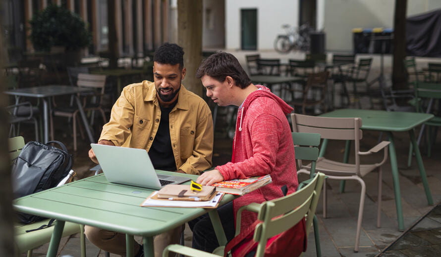 A young man with Down syndrome with his mentoring friend sitting outdoors in cafe using laptop.