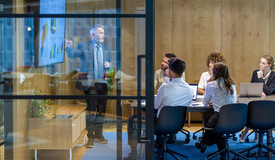 Mature man giving a big data presentation on a tv in a board room. There are several financial graphs and charts on the screen with a diverse group of people in the meeting room. There is paperwork and technology on the table